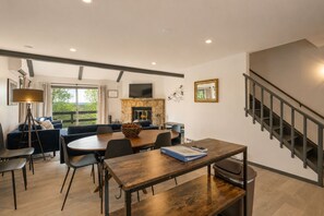 A modern living space with a dining area, a fireplace, a staircase, and a view of greenery.