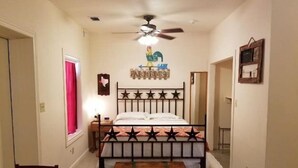 Una habitación estrecha con una cama, un ventilador de techo, un cofre de madera y una mesita.