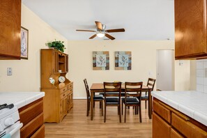 A compact kitchen with wooden cabinets, a dining table with chairs, a ceiling fan, and artwork on the walls.