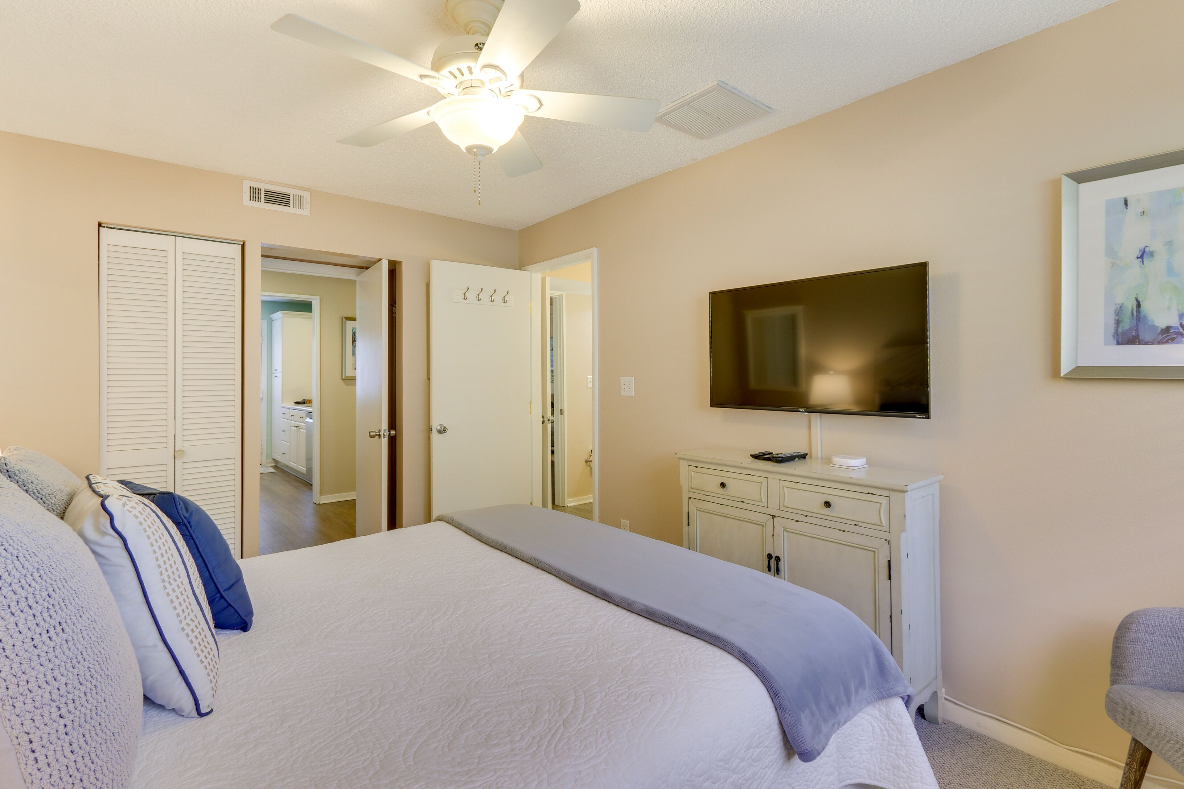Une chambre à coucher comprenant un lit, un ventilateur de plafond, un téléviseur, une commode et une porte donnant sur une autre pièce.