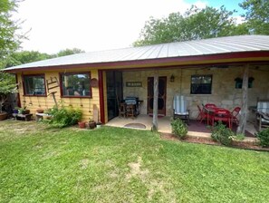 Relax on the covered porch with a table and two chairs, surrounded by a well-maintained lawn and potted plants.