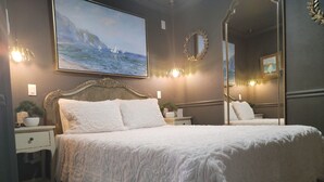 A bedroom with a large bed, a nightstand with a lamp, a wall-mounted mirror, and a painting of a seascape. - Historic Intimate Studio A (Haines City)
