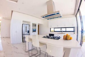 A modern kitchen with white cabinets, a central island with bar stools, a built-in oven, and a large window providing natural light. - Beautiful Ocean And Mountain View  (Las Terrenas)
