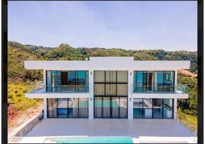 Una casa moderna de dos pisos con amplios ventanales y techo plano, rodeada de vegetación.