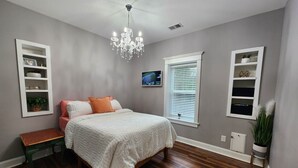 A bedroom with a wooden floor, a bed with white bedding, a bench, a chandelier, and built-in shelving with decorative items. - Barndominium with Access to Horse Farm and Fishing (Comer)