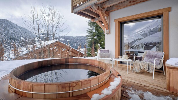 Een houten jacuzzi op een terras met uitzicht op besneeuwde bergen.