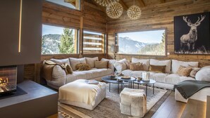 Een gezellig chaletinterieur met een open haard, grote ramen met uitzicht op de bergen en een moderne kroonluchter.