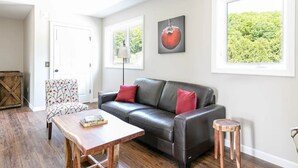 Un salon comprenant un canapé en cuir, une table basse en bois, un fauteuil à motifs et un petit tabouret. Il y a deux fenêtres donnant sur la verdure et une porte menant à une autre pièce.