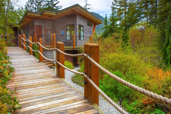 A wooden bridge leading to a cabin with a balcony, surrounded by lush greenery.