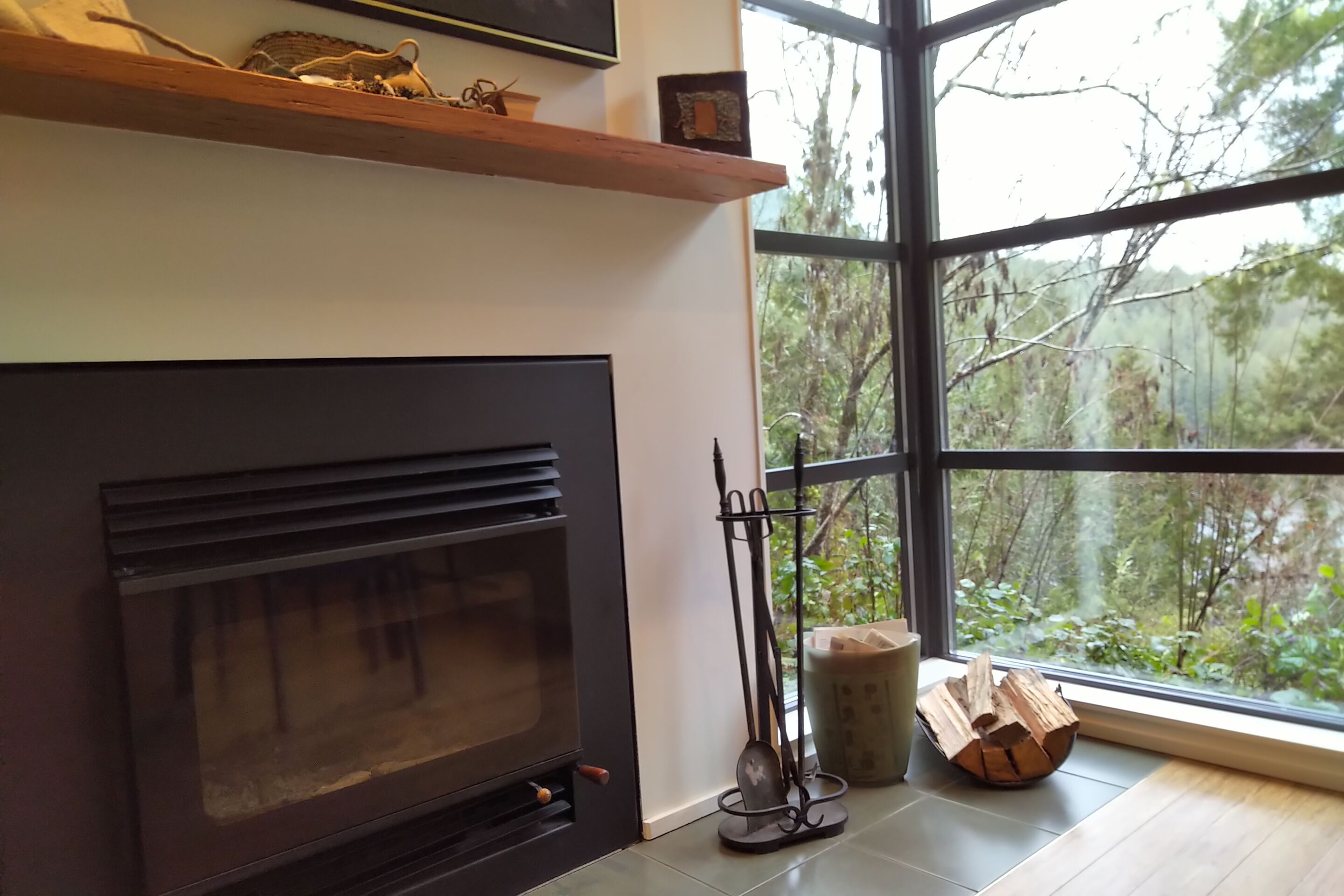 A modern interior with a fireplace, a large window with a view of trees, and a wooden shelf with decorative items.