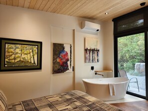 A modern bathroom with a freestanding bathtub, a framed painting, and a large window with a view of trees. - Modern Forest Guest Suite - Accessible Seclusion! (Madeira Park)