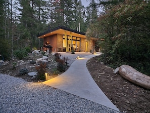 A modern house with a stone exterior, a gravel pathway leading to the entrance, and a forested backdrop.
