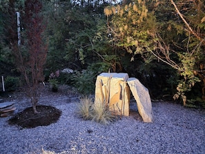 Una escultura de piedra en un jardín con grava, rodeada de árboles y arbustos.