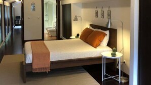 Een moderne hotelkamer met een groot bed, nachtkastje en een bijzettafeltje. Er is een spiegel aan de muur, een klein plantje op het nachtkastje, en een raam dat natuurlijk licht biedt.