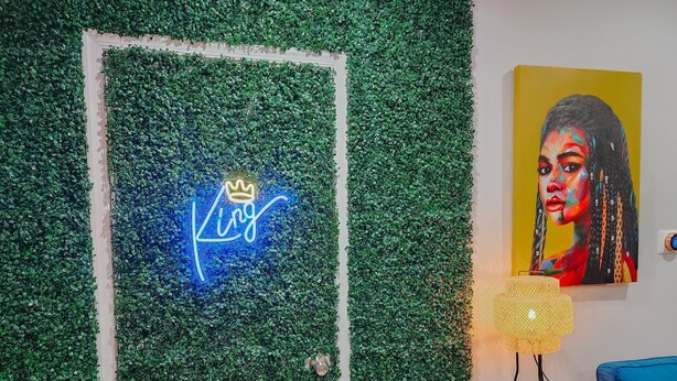 Ein Zimmer mit grüner Wand, einem Neon-Schild mit der Aufschrift 'King' und einer Krone, einem Bild einer Frau und einem blauen Stuhl.