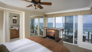 Een slaapkamer met een groot bed, een plafondventilator, een open haard en een balkon met uitzicht op de oceaan.