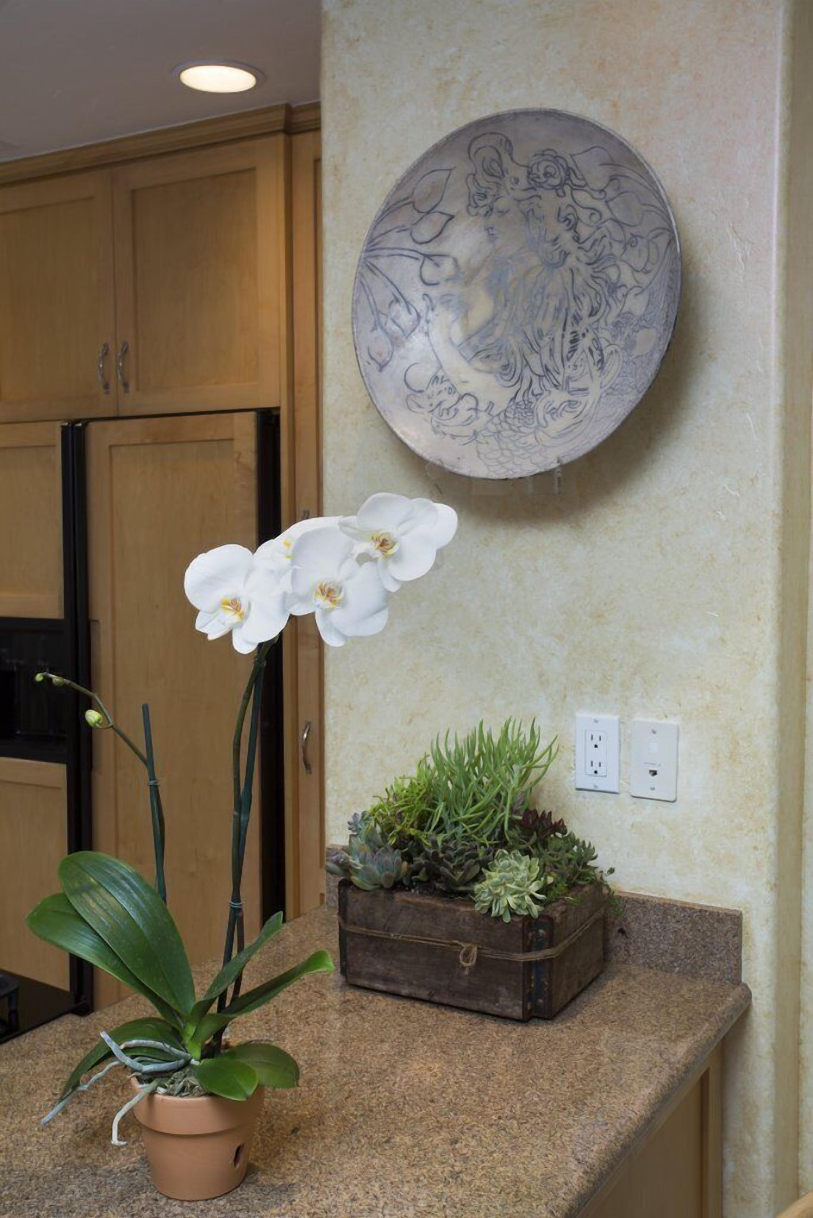A kitchen with wooden cabinets, a potted orchid, a small plant in a vintage box, and a decorative plate on the wall.