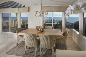 Ein Essbereich mit großem Tisch, Korbstühlen, einer Chandelierleuchtre und Blick auf den Ozean durch große Fenster.