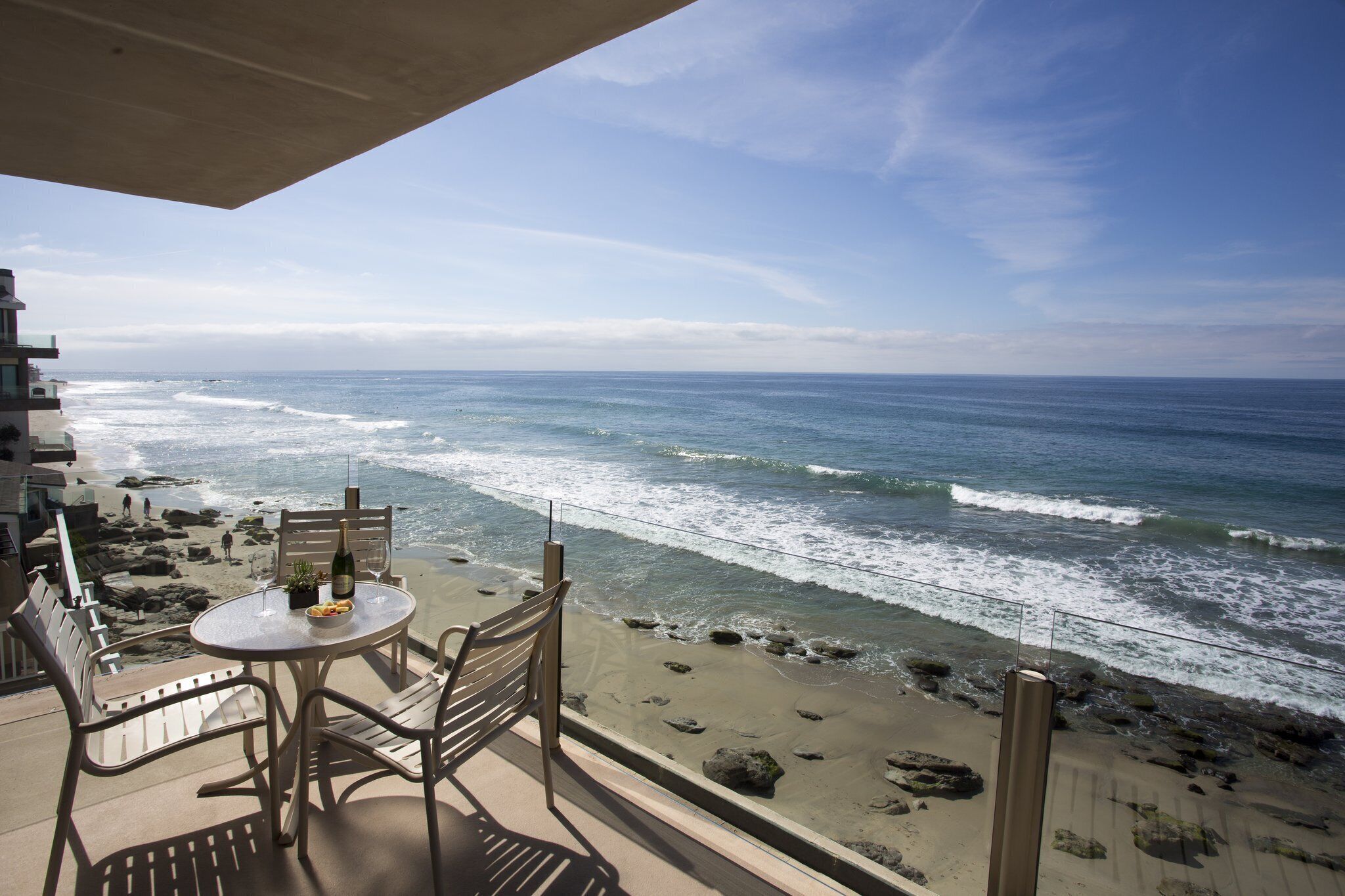 Ein Balkon mit Tisch, Stühlen und einer Weinflasche, mit Blick auf einen Strand mit Wellen und Felsen.