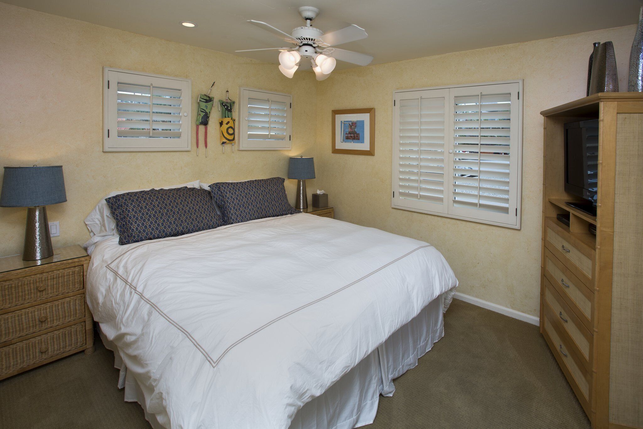 A bedroom with a large bed, two bedside tables with lamps, a ceiling fan, and a wooden dresser with a television.
