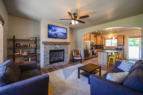 A spacious living room with a fireplace, a television mounted above it, and a modern kitchen in the background. - Coeur d'Alene Hideaway 4 Bedroom Modern Mid Term Rental (Coeur d'Alene)