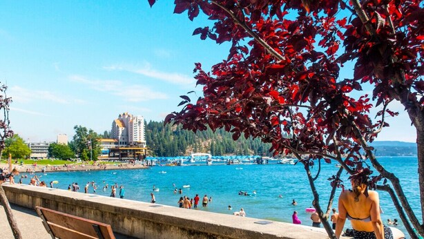 A waterfront promenade with a bench, a tree with red leaves, and a view of a beach with people swimming and sunbathing. - Coeur d'Alene Hideaway 4 Bedroom Modern Mid Term Rental (Coeur d'Alene)