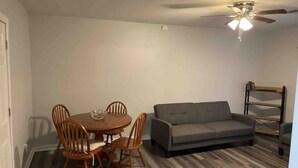 Une pièce comprenant une table à manger, des chaises, un canapé gris, un ventilateur de plafond et une étagère en bois.