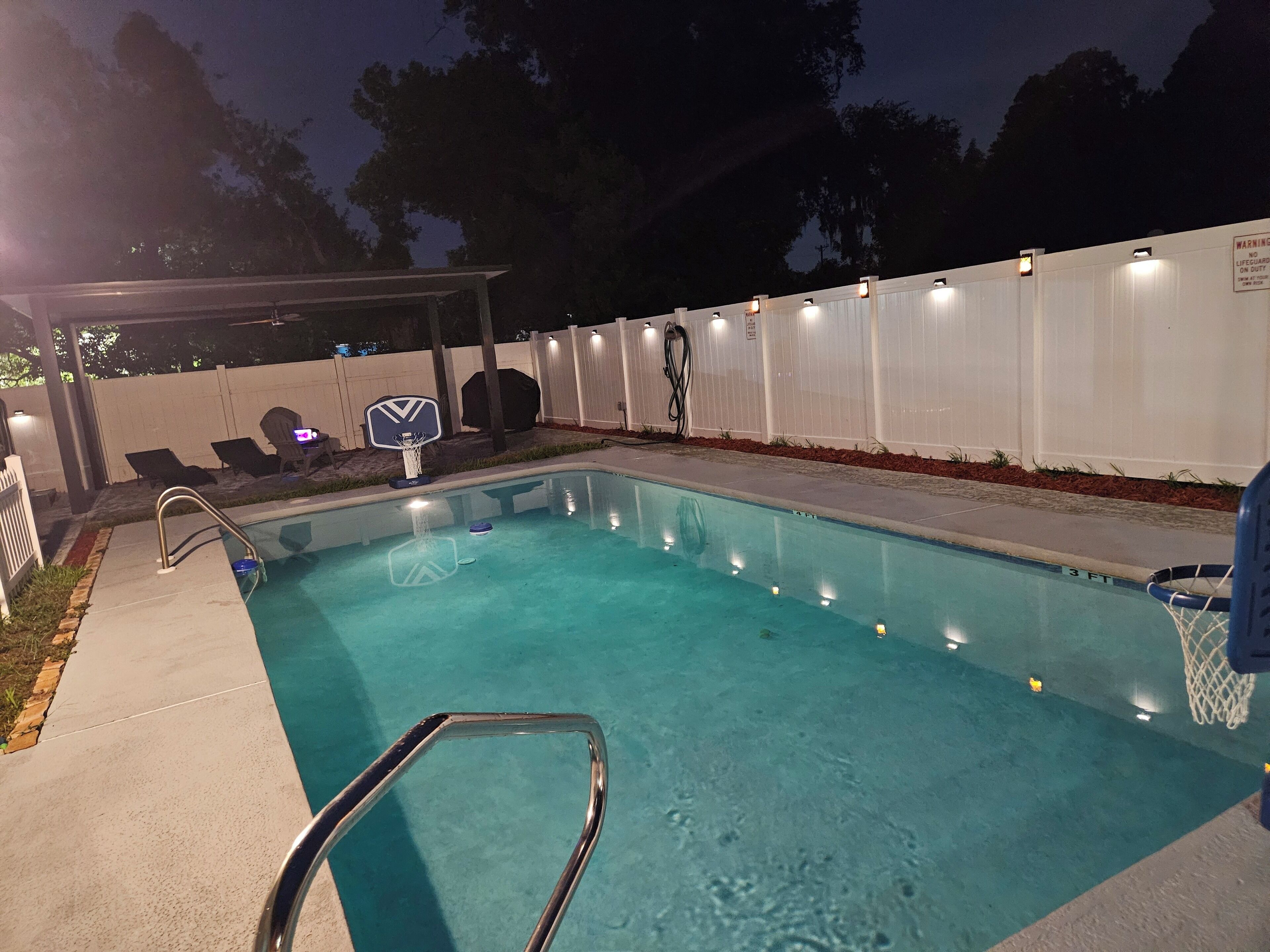 A swimming pool with a basketball hoop, a covered seating area, and a fenced enclosure with lights.