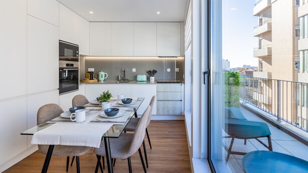 Une cuisine moderne avec des meubles blancs, un coin repas doté d’une table en verre et un balcon offrant une vue sur la ville.