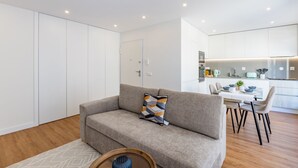 Un salon moderne avec un canapé gris, un coussin à motifs et une table d’appoint en bois. On aperçoit à l’arrière-plan un coin repas avec une table et des chaises blanches.