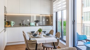 Une cuisine moderne avec des meubles blancs, une table à manger pour quatre personnes et une vue sur l’extérieur grâce à de grandes fenêtres.