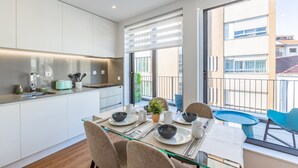 Une cuisine moderne avec une table à manger en verre, des meubles blancs et un balcon agrémenté d’une chaise bleue.
