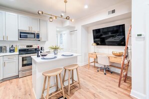 A modern kitchen with white cabinets, a microwave, stove, and a large TV. There are stools at a small island and a desk with a chair. - Lincoln Lofts: 202 | Luxe Boho 2BR in Heart of DC (Washington)