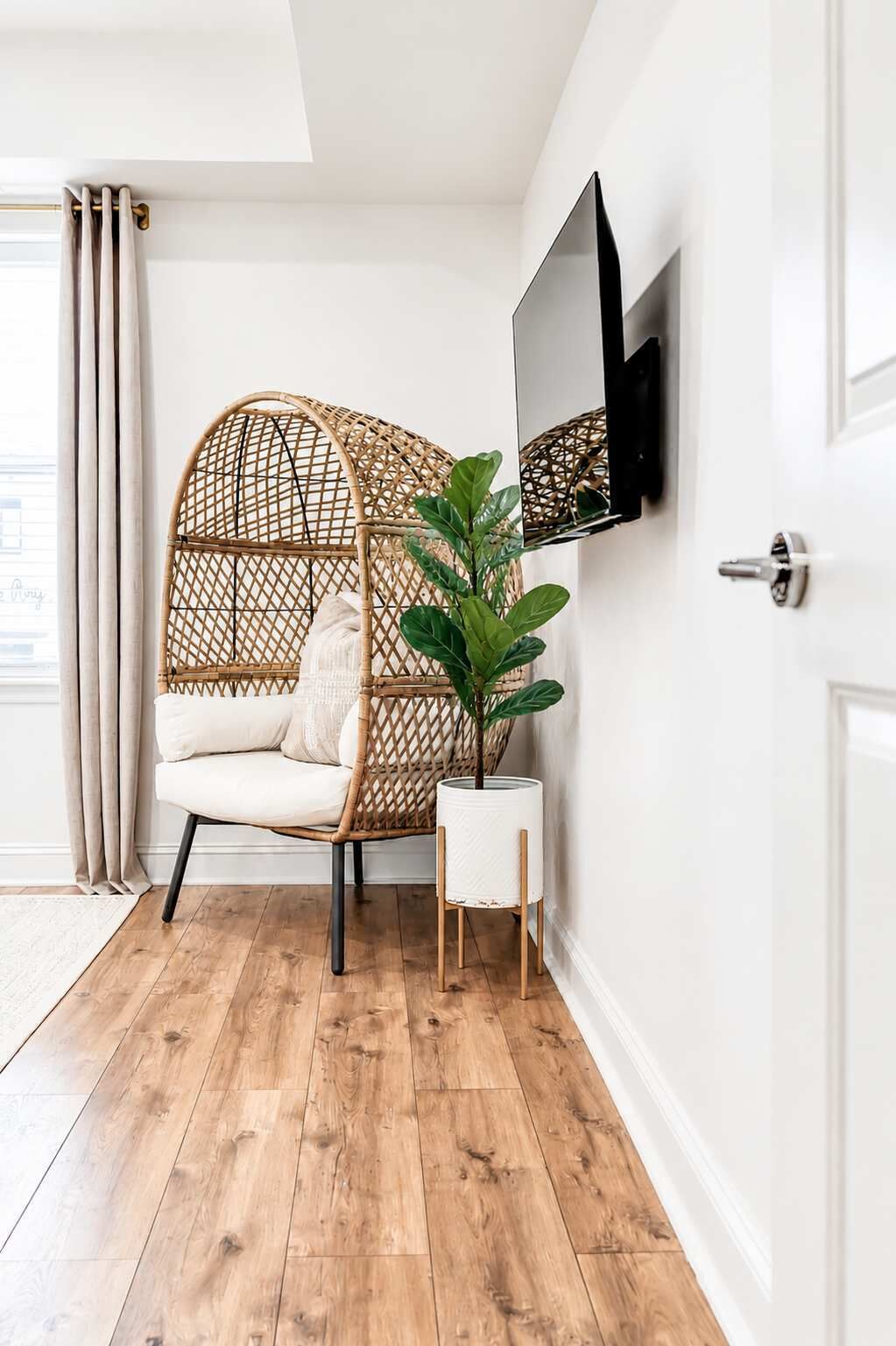 Une pièce avec un plancher en bois, une chaise blanche avec des coussins, une chaise en osier, une plante en pot, un téléviseur fixé au mur et une porte.