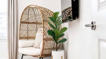 Une pièce avec un plancher en bois, une chaise blanche avec des coussins, une chaise en osier, une plante en pot, un téléviseur fixé au mur et une porte.