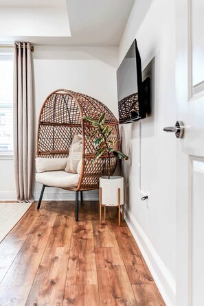 A room with a wooden floor, a wicker chair with cushions, a small white side table with a plant, and a mounted flat-screen TV. - Lincoln Lofts: 202 | Luxe Boho 2BR in Heart of DC (Washington)