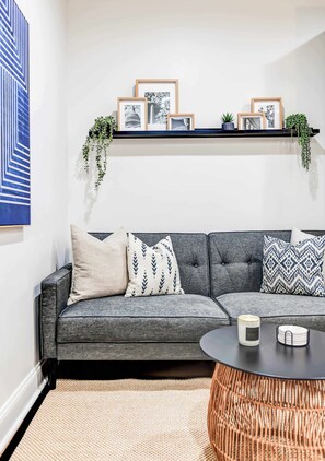Ein modernes Wohnzimmer mit einem grauen Sofa, gemusterten Kissen, einem runden Couchtisch und einem schwarzen Regal mit gerahmten Bildern und Pflanzen.