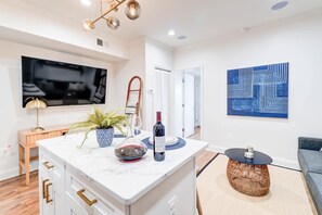 A modern kitchen with a marble countertop, a TV mounted above it, and a dining area with a round table and wicker chair. - Lincoln Lofts: 202 | Luxe Boho 2BR in Heart of DC (Washington)