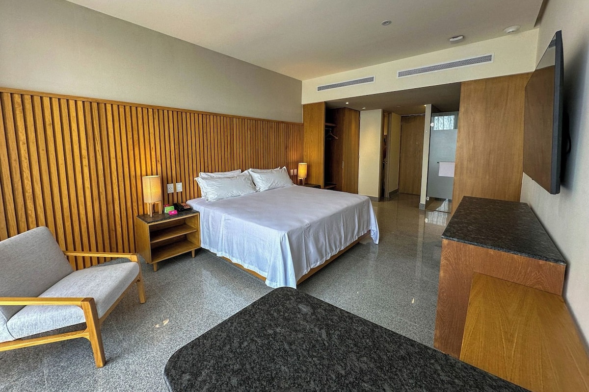 A modern hotel room with a large bed, a wooden headboard, a seating area with a chair, a small table, and a flat-screen TV mounted on the wall.