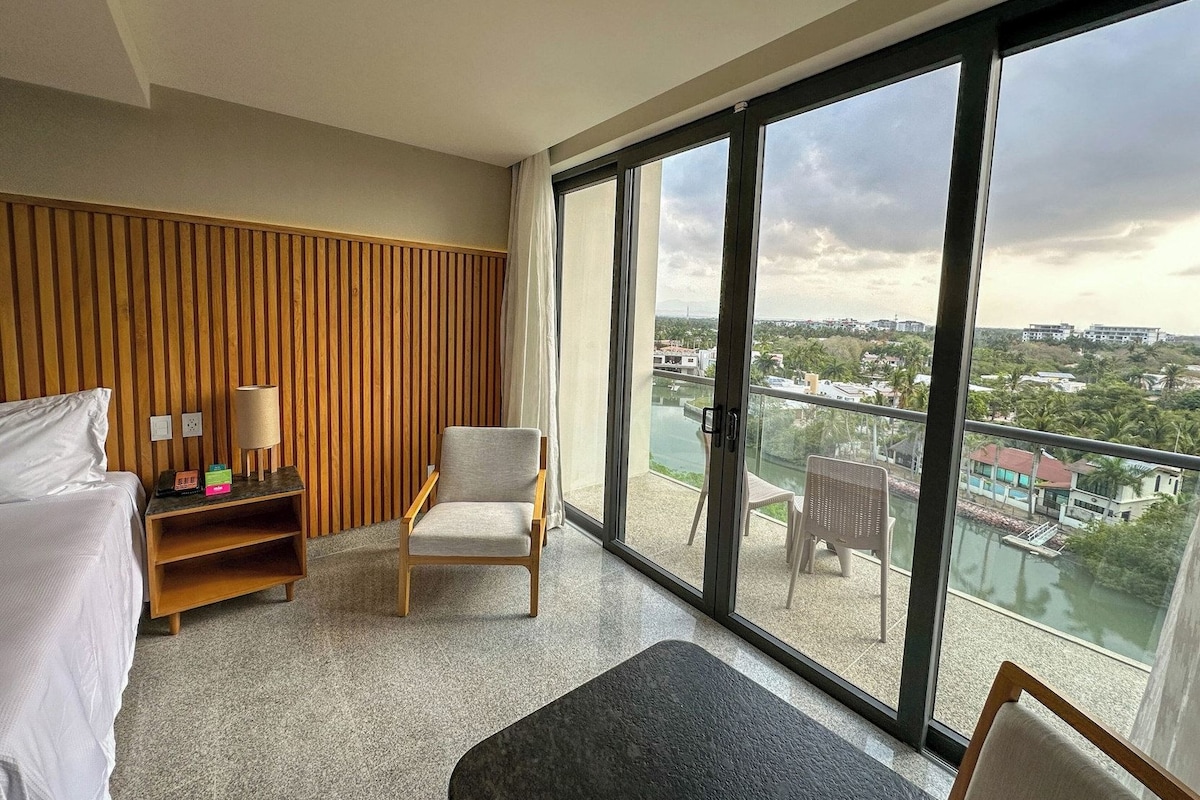 Habitación de hotel con cama, una silla, una mesita con lámpara y un ventanal que ofrece vistas a la ciudad y al agua.