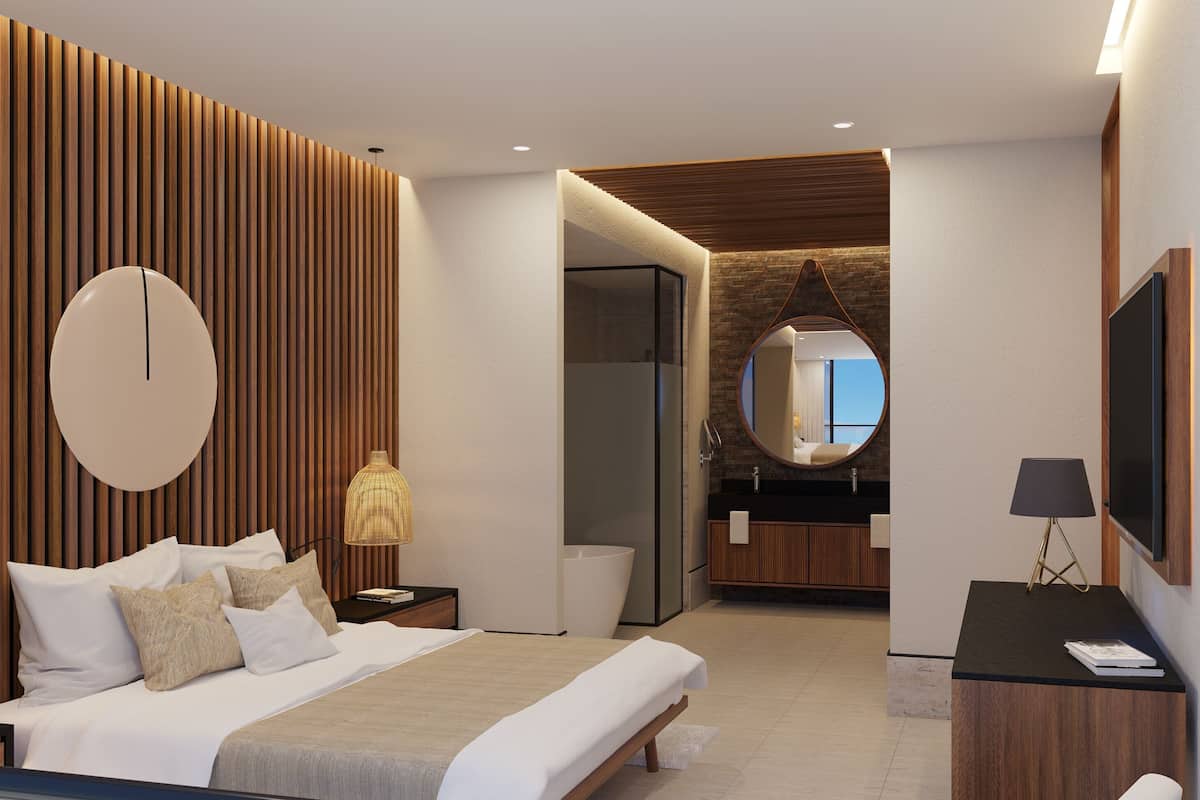 A modern hotel room with a bed, bedside table, lamp, TV, and a bathroom with a mirror and sink.