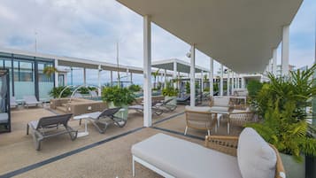 Un área de lounge al aire libre, techada, con diferentes disposiciones de asientos, incluyendo sillones y sofás, y plantas en macetas.