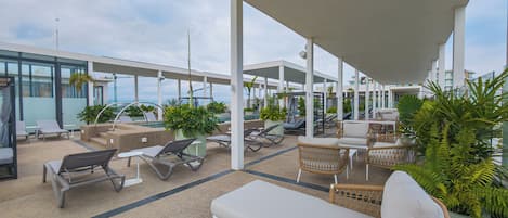 Un área de lounge al aire libre, techada, con diferentes disposiciones de asientos, incluyendo sillones y sofás, y plantas en macetas.