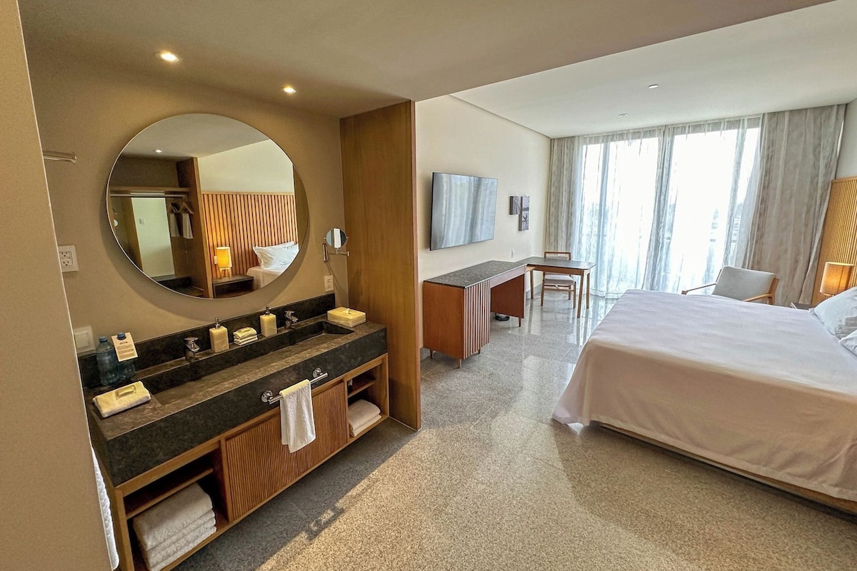 A hotel room with a large bed, a desk, a chair, a TV, and a bathroom with a sink and mirror.