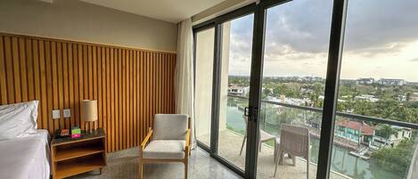 Habitación de hotel con cama, una silla, una mesita de noche con lámpara y un ventanal que ofrece vistas de un río y edificios.