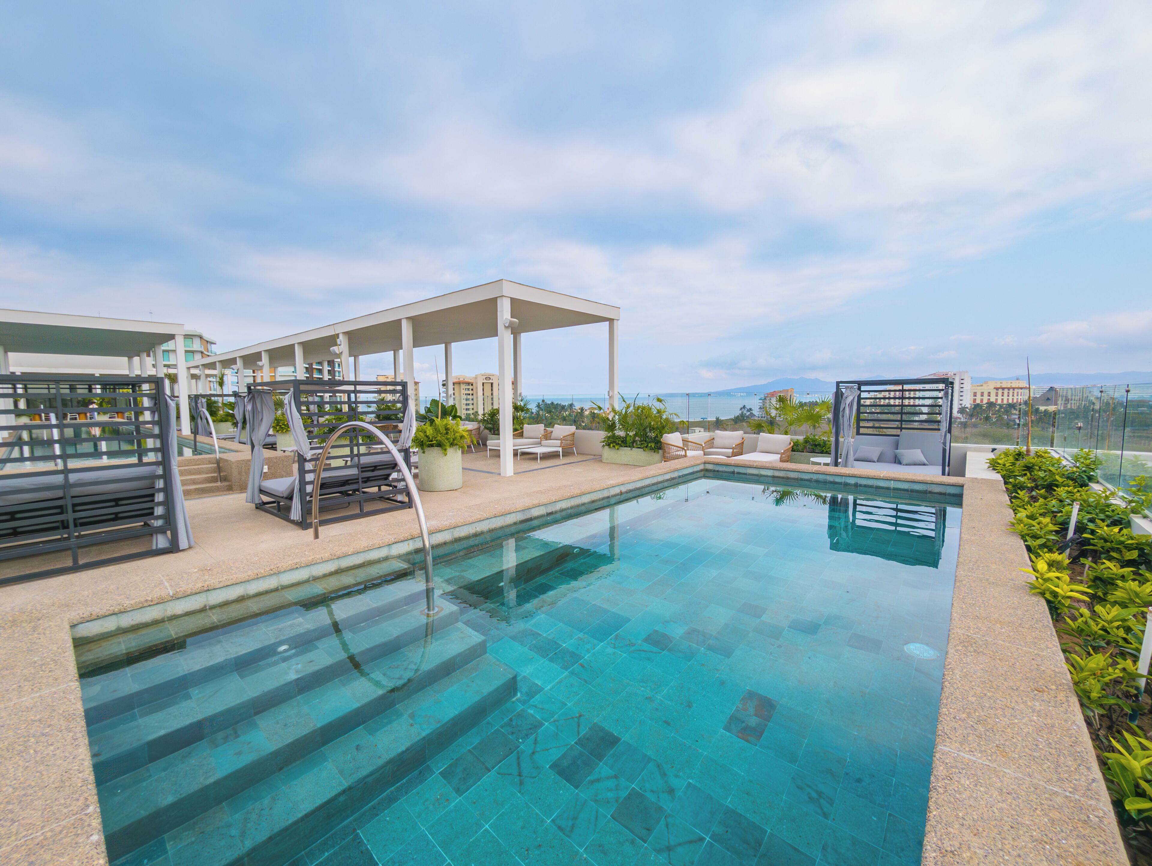 Une piscine à débordement située sur le toit, avec des chaises longues et un espace salon couvert, offrant une vue panoramique sur la ville.