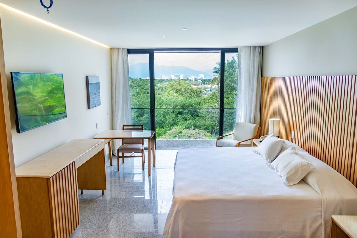 Habitación de hotel con cama, escritorio, silla, televisor y un amplio ventanal con vistas a zonas verdes y edificios.