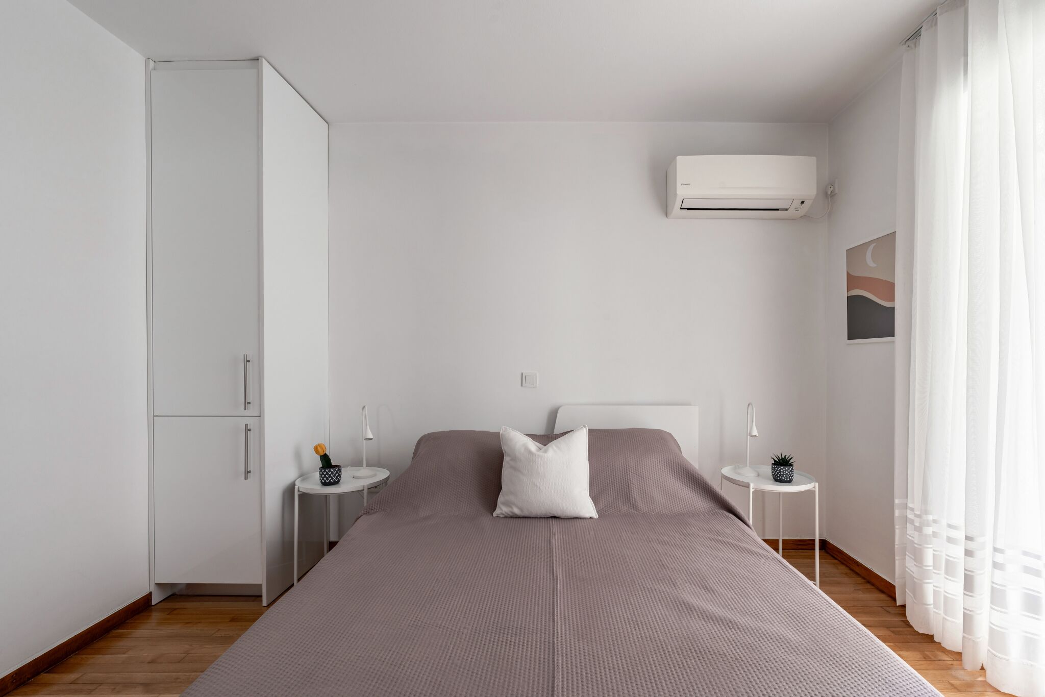 Een minimalistische slaapkamer met een enkel bed, witte muren, een kledingkast, een klein nachtkastje met een lamp en een wandairconditioner.