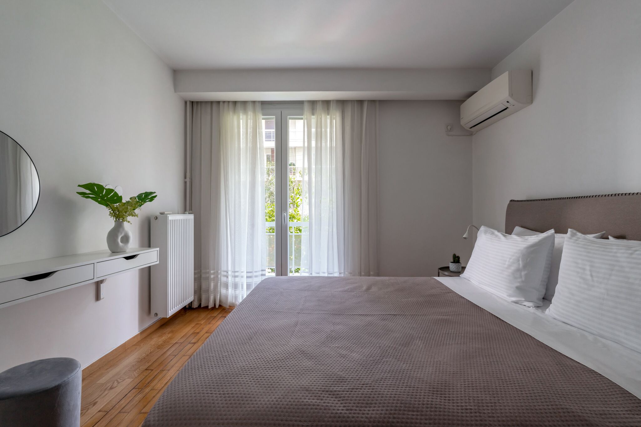 Une chambre moderne avec un grand lit, une table de chevet, une plante en pot et une vue sur l’extérieur à travers des rideaux légers.