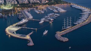 An aerial view of a marina with multiple yachts docked, a breakwater, and a cityscape in the background. - The Black Marble - Sea View / 2BR / 1,5BA (Paleo Faliro)